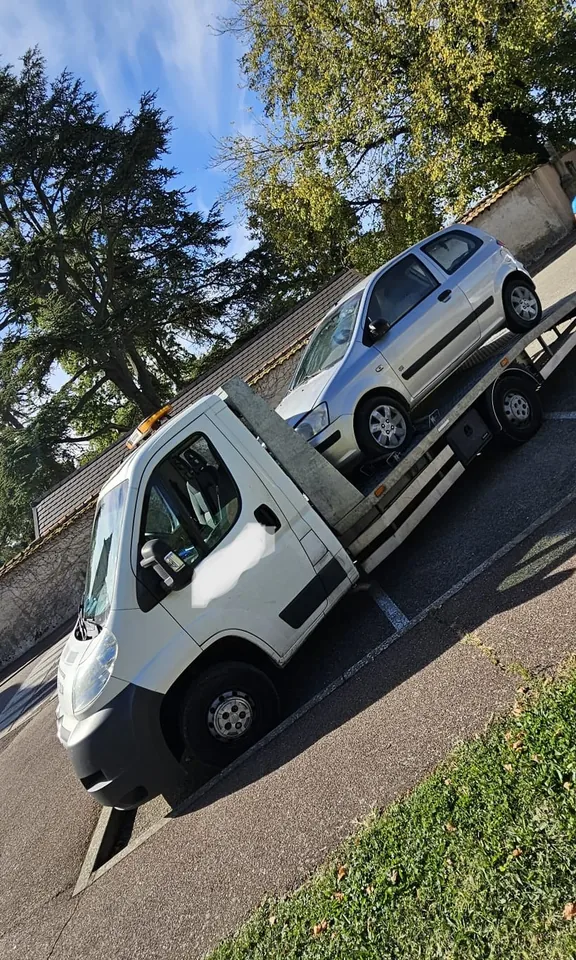 Citadine à trois portes sur un camion-plateau près d’un mur et d’arbres.