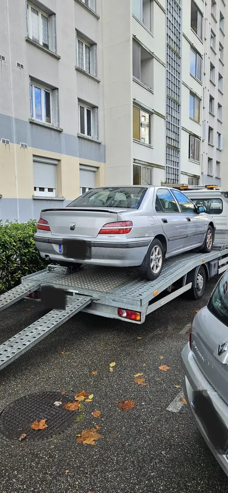 Voiture grise sur un camion plateau avec rampe abaissée devant des immeubles.