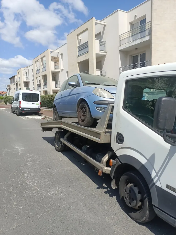 Voiture sombre sur le plateau d’un camion blanc dans une rue bordée d’immeubles.