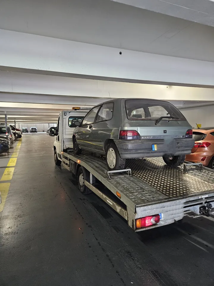Petite voiture grise chargée sur un camion plateau dans un parking couvert.