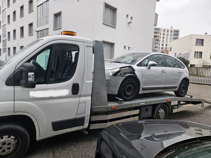 Voiture claire au capot et à l’avant déformés sur un camion devant des immeubles.