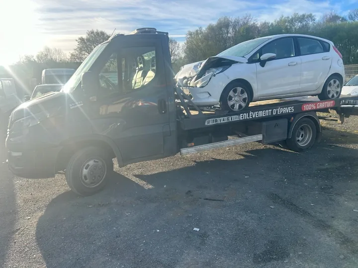 Petite voiture claire installée sur un camion plateau sombre en extérieur.