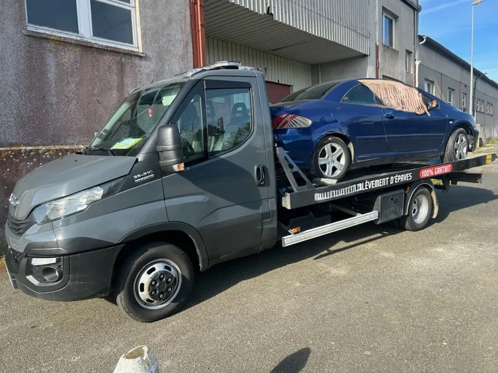 Voiture bleue sur le plateau d’un camion gris, devant un bâtiment.