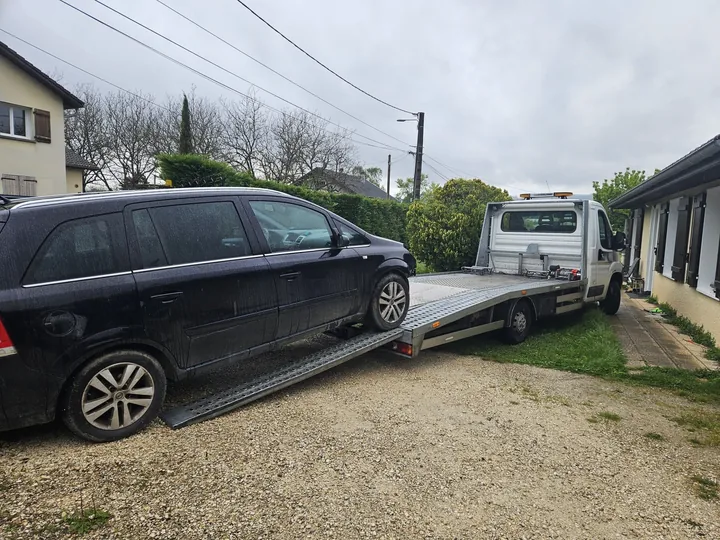Voiture sombre près d’un camion plateau dans une cour bordée de bâtiments.