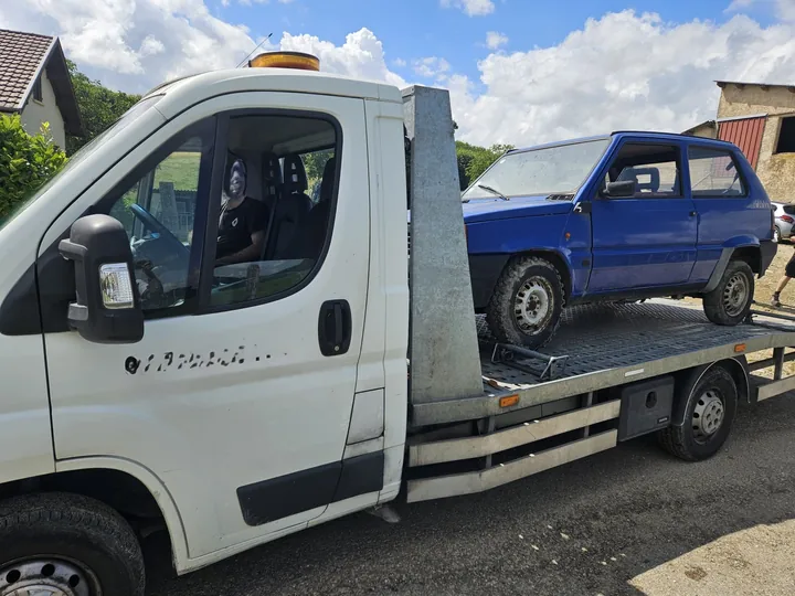 Véhicule bleu avec une benne arrière installé sur le plateau d’un camion blanc.