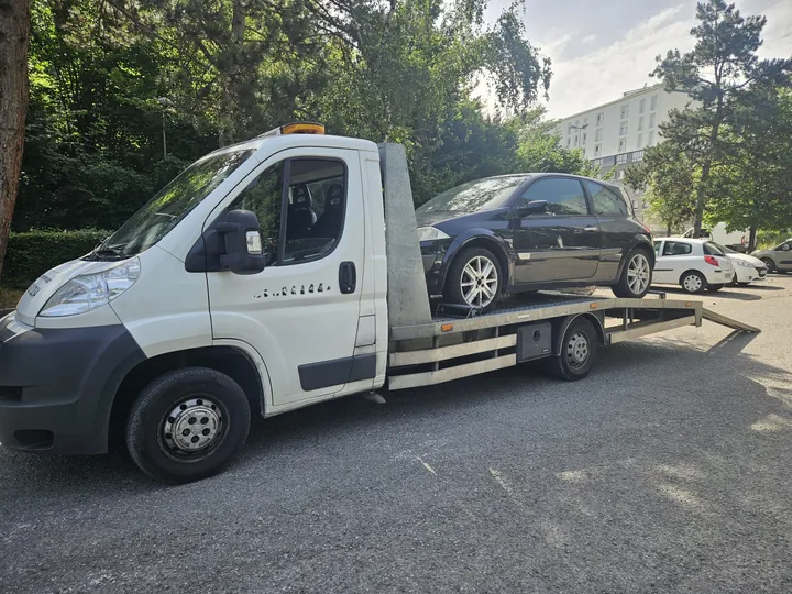 Camion blanc portant une voiture grise sur son plateau près d’arbres.