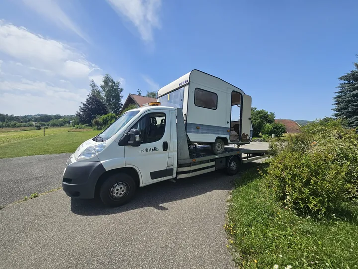 Dépanneuse blanche transportant un petit véhicule à cellule habitable en bord de route.