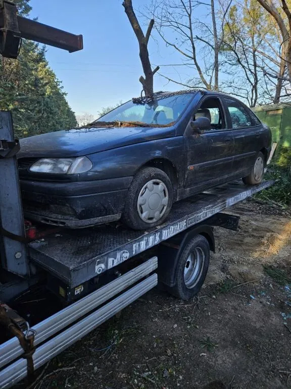 Voiture sombre chargée sur un plateau, près d’arbres et d’une zone clôturée.