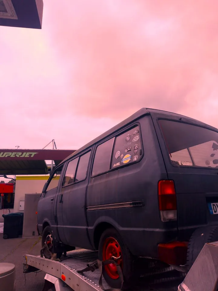 Ancien fourgon sombre sur une dépanneuse sous un ciel teinté de rose.