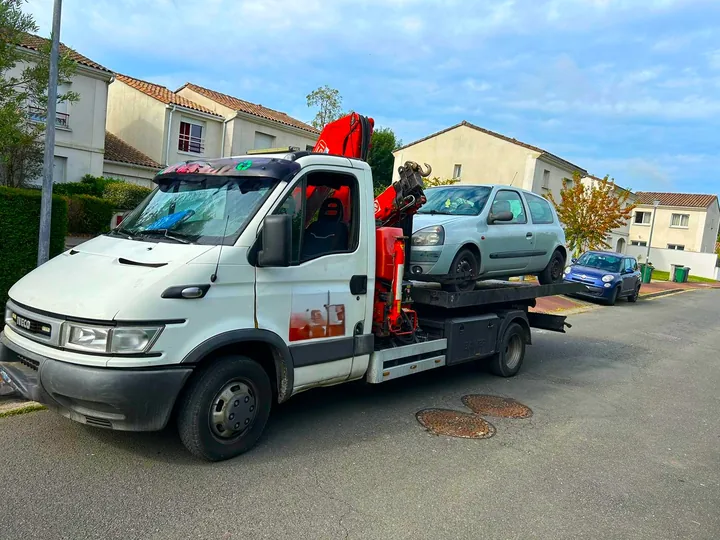 Dépanneuse à grue repliée portant une citadine claire dans une rue.