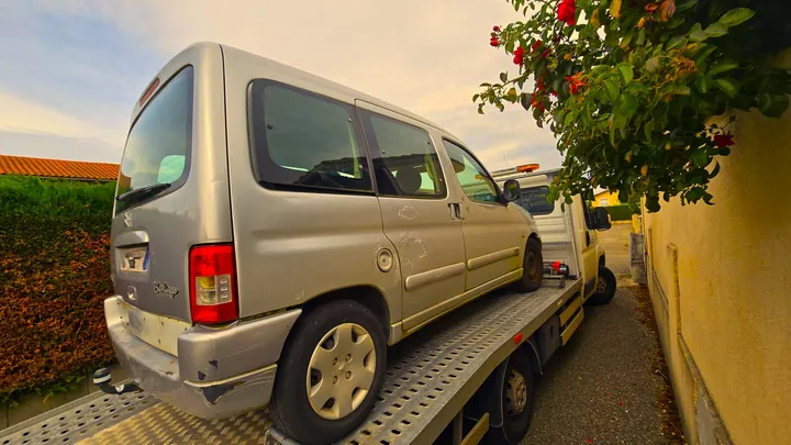 Petite voiture gris clair sur une dépanneuse près d’un mur et d’un arbre.