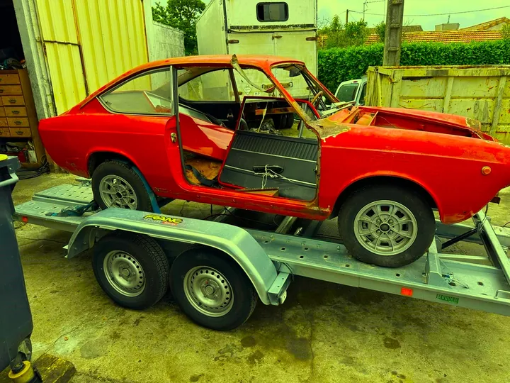 Voiture ancienne sans capot ni pare-brise, avec une portière ouverte, sur une remorque.